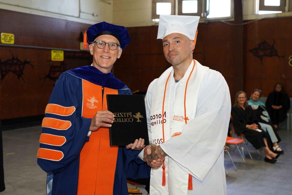 里维拉穿着白色和橙色的徽章，塞克穿着深蓝色和橙色的徽章. 里维拉和塞克握手，塞克递给他一张推荐十大正规网赌网站的黑人毕业证书.