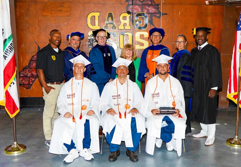 三位穿着白色和橙色徽章的Inside-Out毕业生坐在前面. 一群七人站在他们身后，穿着不同颜色的毕业典礼礼服.